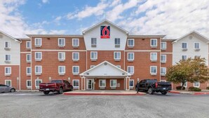 A 6 Suites hotel with a brick facade, a central entrance, and a parking lot with two vehicles. - 2 Full Beds | Smoking, Kitchen (Oklahoma City OK)