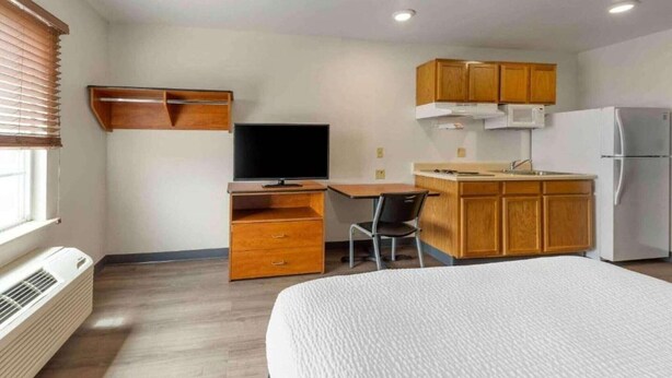 Habitación de hotel con cama, escritorio con silla, televisor, kitchenette equipada con microondas y refrigerador, y ventana con persianas.
