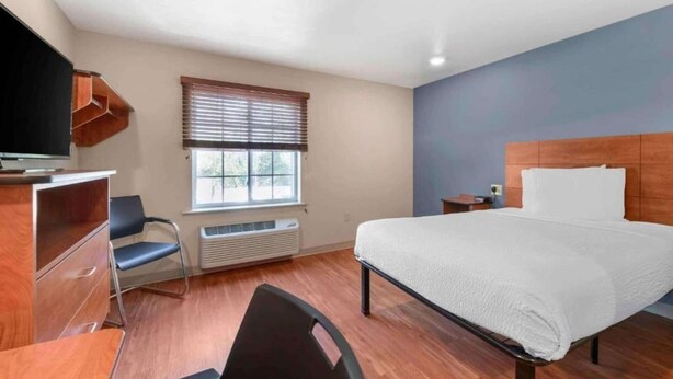A hotel room with a bed, a desk, a chair, a television, a window with blinds, and a heater.