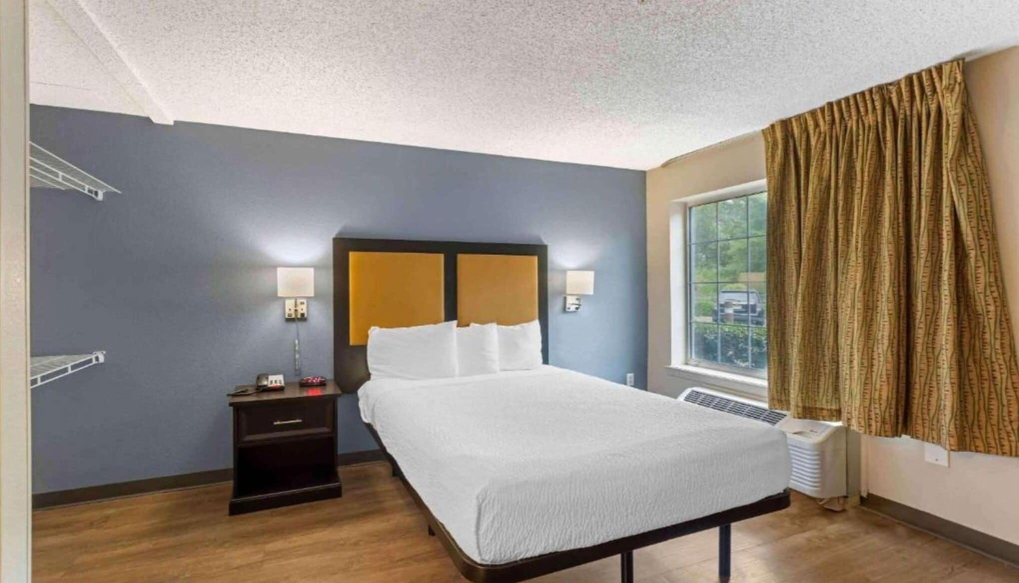 A hotel room with a bed, bedside table, lamp, window with curtains, and a view of the outside.