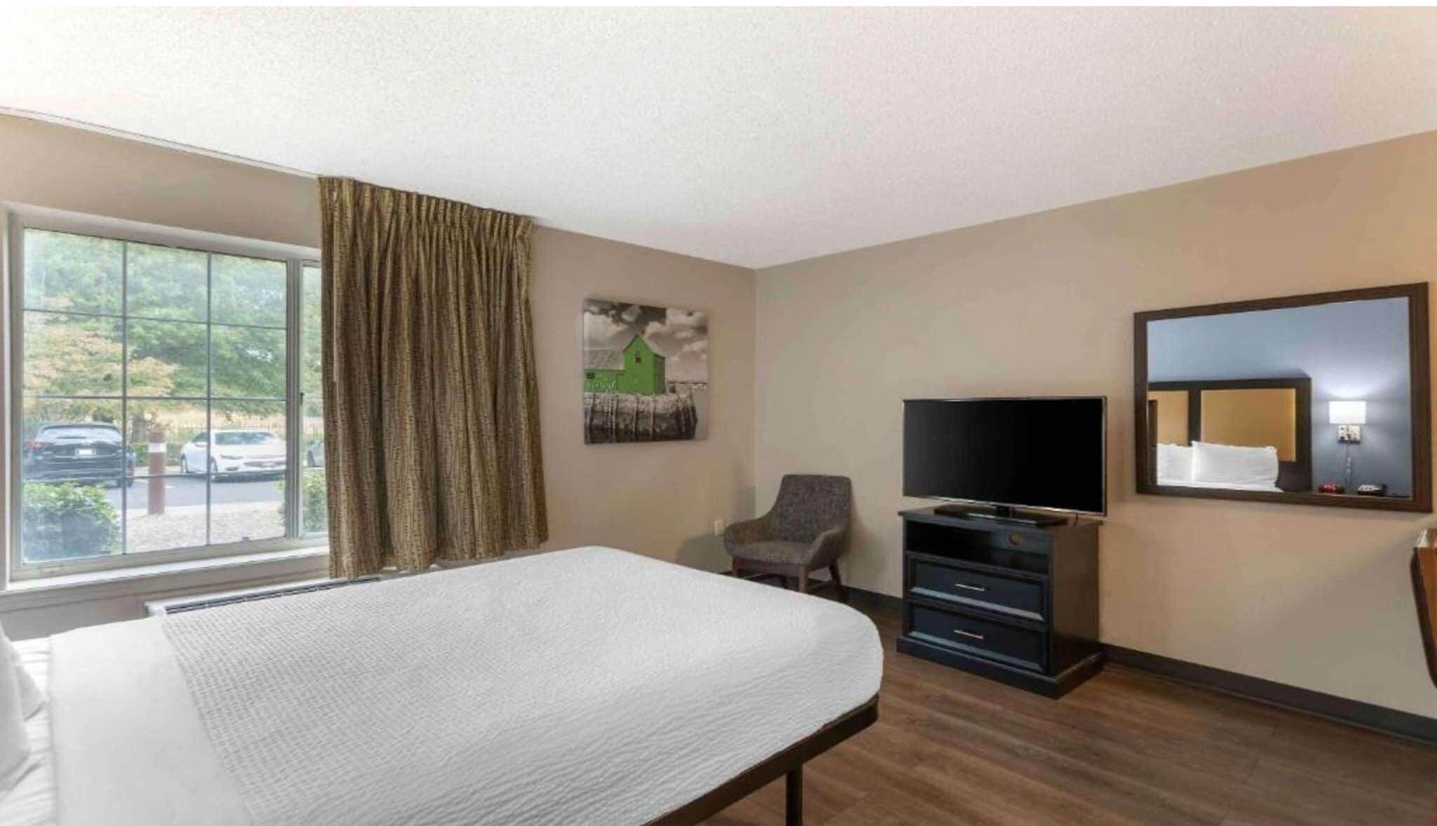 Una habitación de hotel con cama, televisión, silla, cómoda y una ventana grande con cortinas.
