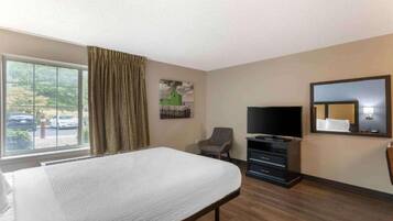 Una habitación de hotel con cama, televisión, silla, cómoda y una ventana grande con cortinas.