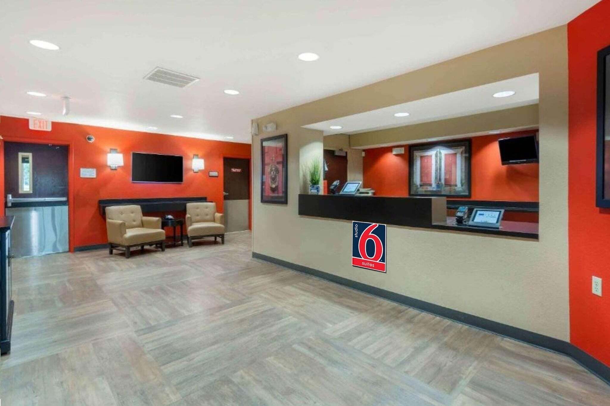 A hotel reception area with a reception desk, two armchairs, a small table, and a TV screen.