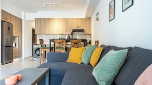 Een moderne woonkamer met een grijze bank, een salontafel en een eetgedeelte met houten kasten en een keuken op de achtergrond.