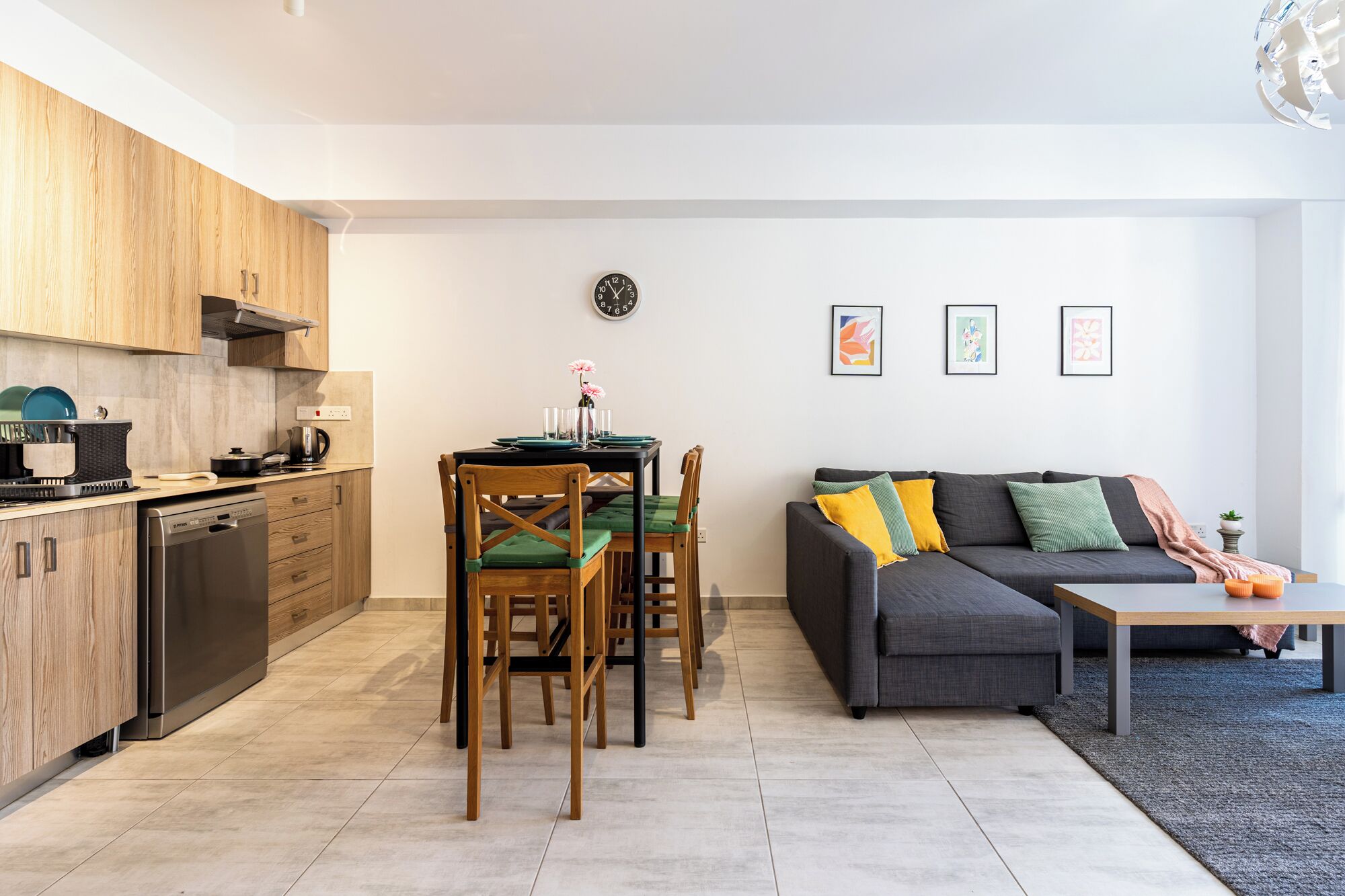 A modern kitchen with wooden cabinets, a dining area with a table and chairs, a gray sofa with cushions, and a small side table.
