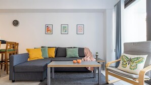 Een moderne woonkamer met een grijze bank, een houten stoel, een salontafel en drie kunstwerken in lijsten aan de muur.