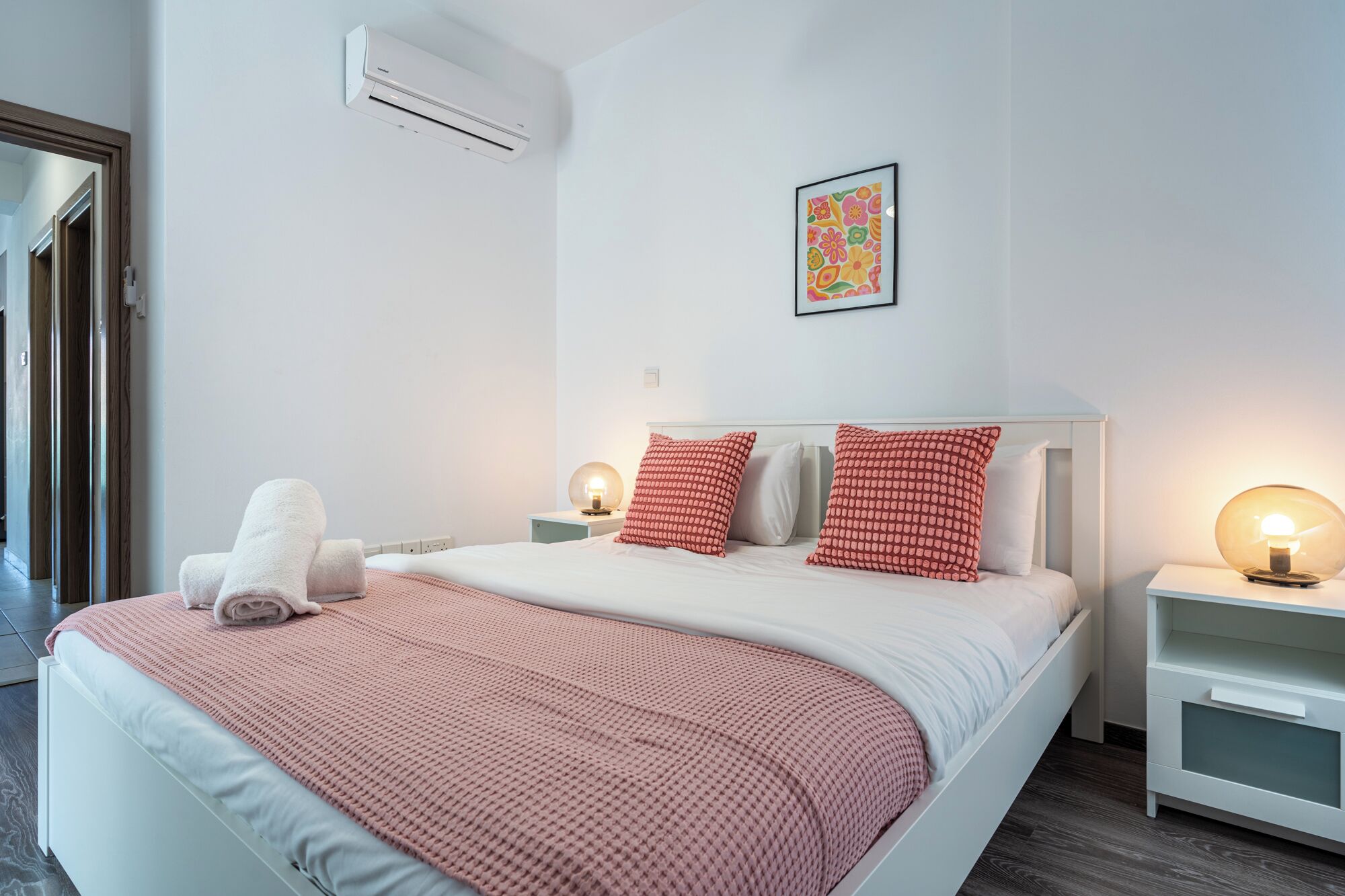 Un dormitorio con cama, almohadas, una mesita de noche con lámpara, equipo de aire acondicionado y un cuadro en la pared.