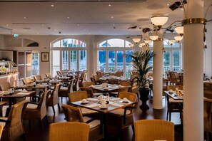 Un restaurante espacioso con varias mesas y sillas, ventanas amplias y un moderno candelabro.