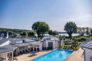 A swimming pool with lounge chairs and umbrellas, surrounded by buildings with metal roofs and a view of a lake. - Schwielowsee  Hotel Superior Double Room (Werder)