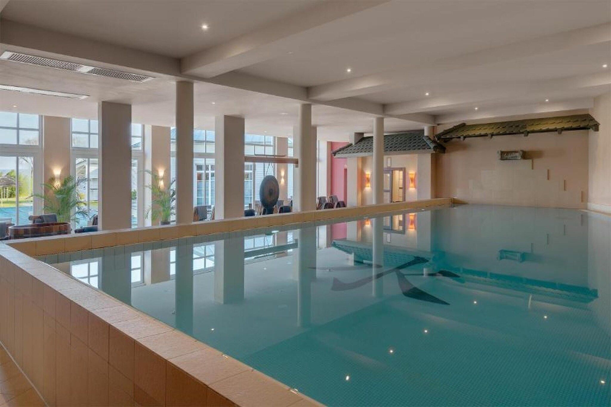 Una piscina interior con agua cristalina, grandes ventanales y suelo de baldosas.