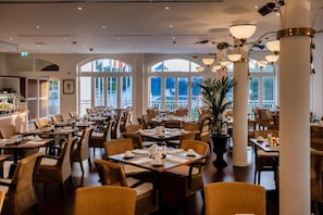Een restaurant met meerdere eettafels en stoelen, grote ramen en een centrale pilaar.