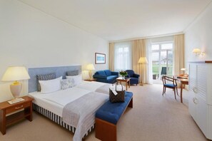 Een hotelkamer met een groot bed, twee blauwe fauteuils, een bureau met stoel en uitzicht op het landschap door het raam.