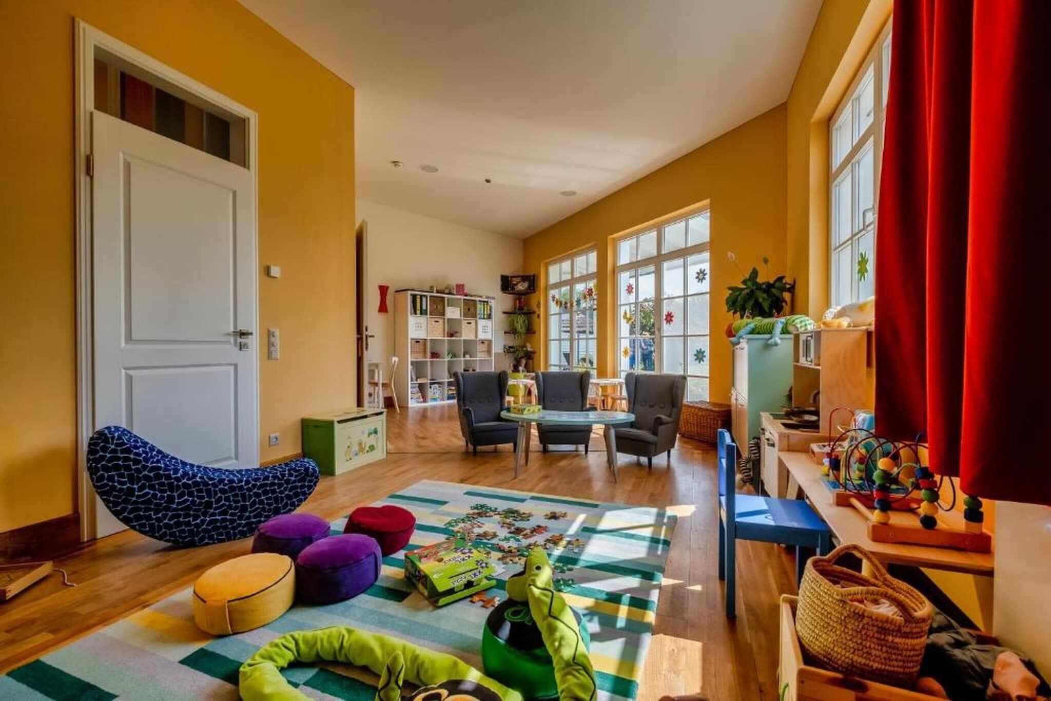 Una habitación con piso de madera, una puerta blanca, un sillón de peluche, una pequeña mesa con sillas, una estantería y una ventana con cortinas.