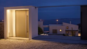 A modern house with a well-lit entrance, a driveway, and a view of the sea at dusk. - NISIS II – Urban Loft Surrounded by Nature (Perani)