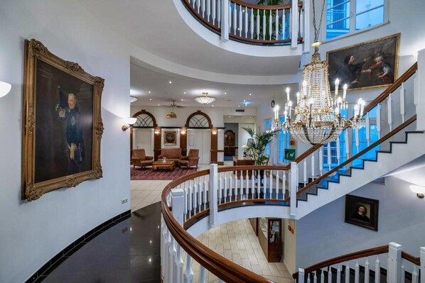 Un intérieur somptueux avec un escalier, des lustres et des œuvres d’art encadrées.