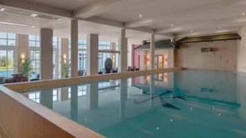 Una piscina interior con agua cristalina, grandes ventanales y suelo de baldosas.