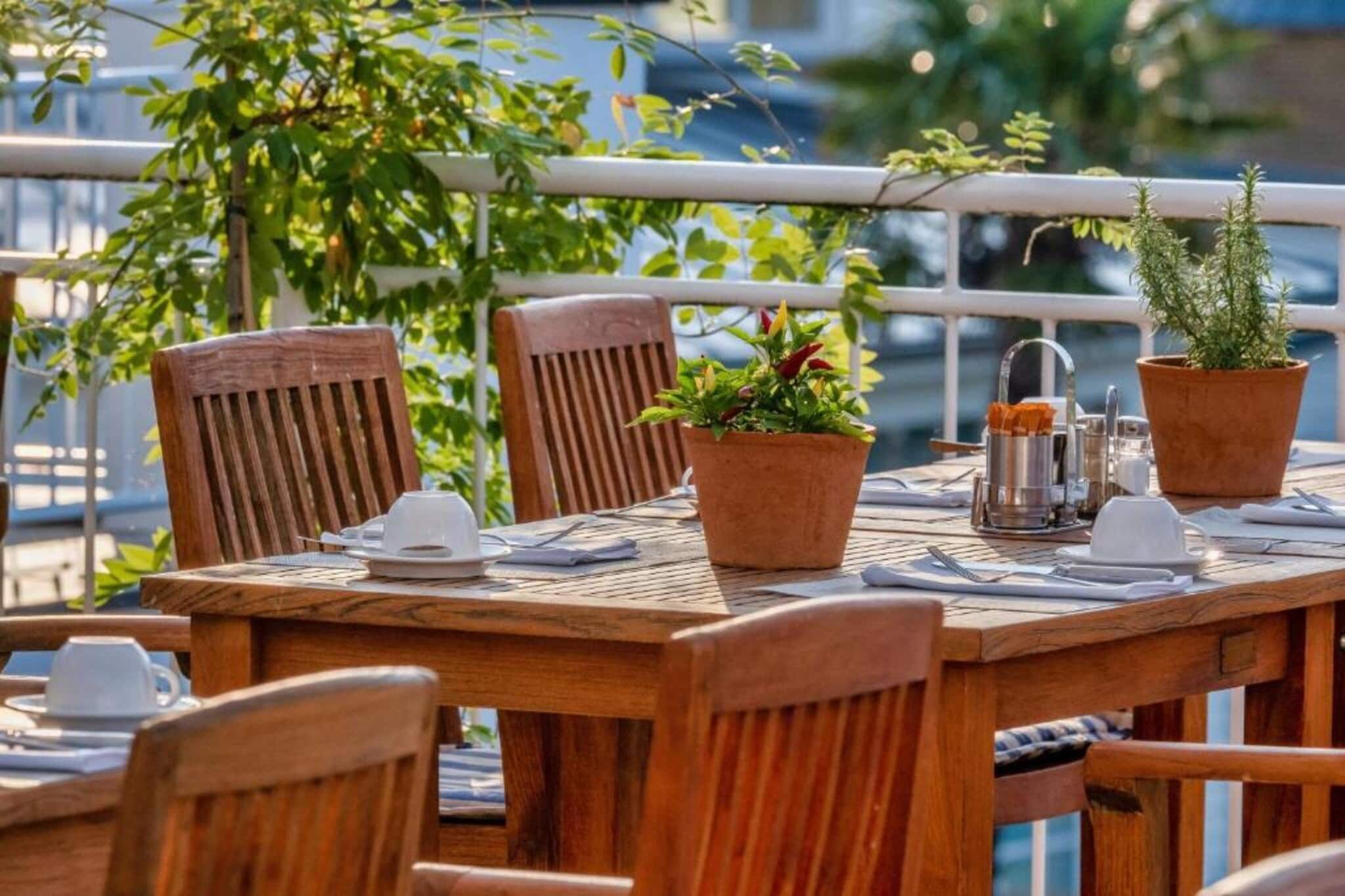 Mesa de madeira ao ar livre com cadeiras, plantas em vasos e uma cafeteira de metal.