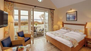 A bedroom with a bed, two chairs, a table with fruit, a TV, and a balcony with a view.