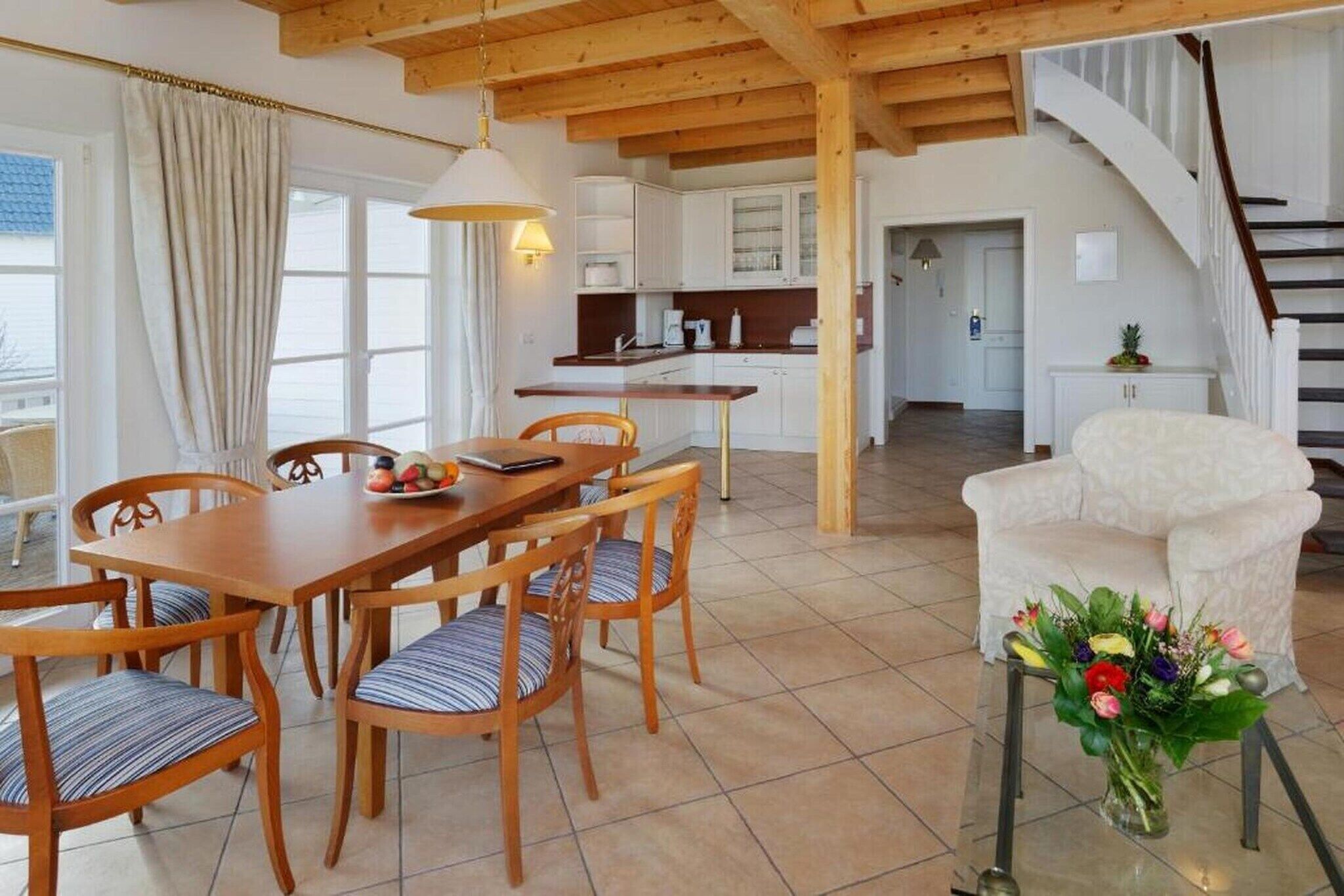Un coin repas avec une table et des chaises en bois, une cuisine à l’arrière-plan et un escalier menant à un autre niveau.