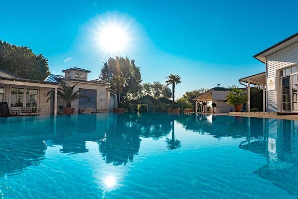 Une piscine aux eaux cristallines, baignée de lumière, avec un ciel ensoleillé, et entourée de maisons et d’arbres.