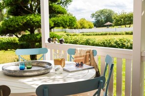 Une table avec un plateau, des verres et une carafe, placée sur une table blanche dans un espace extérieur abrité, avec des chaises bleues et une vue sur un jardin.