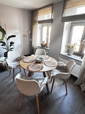 Un coin repas avec une table ronde, des chaises blanches, des sets de table en fibres tressées et une plante en pot.