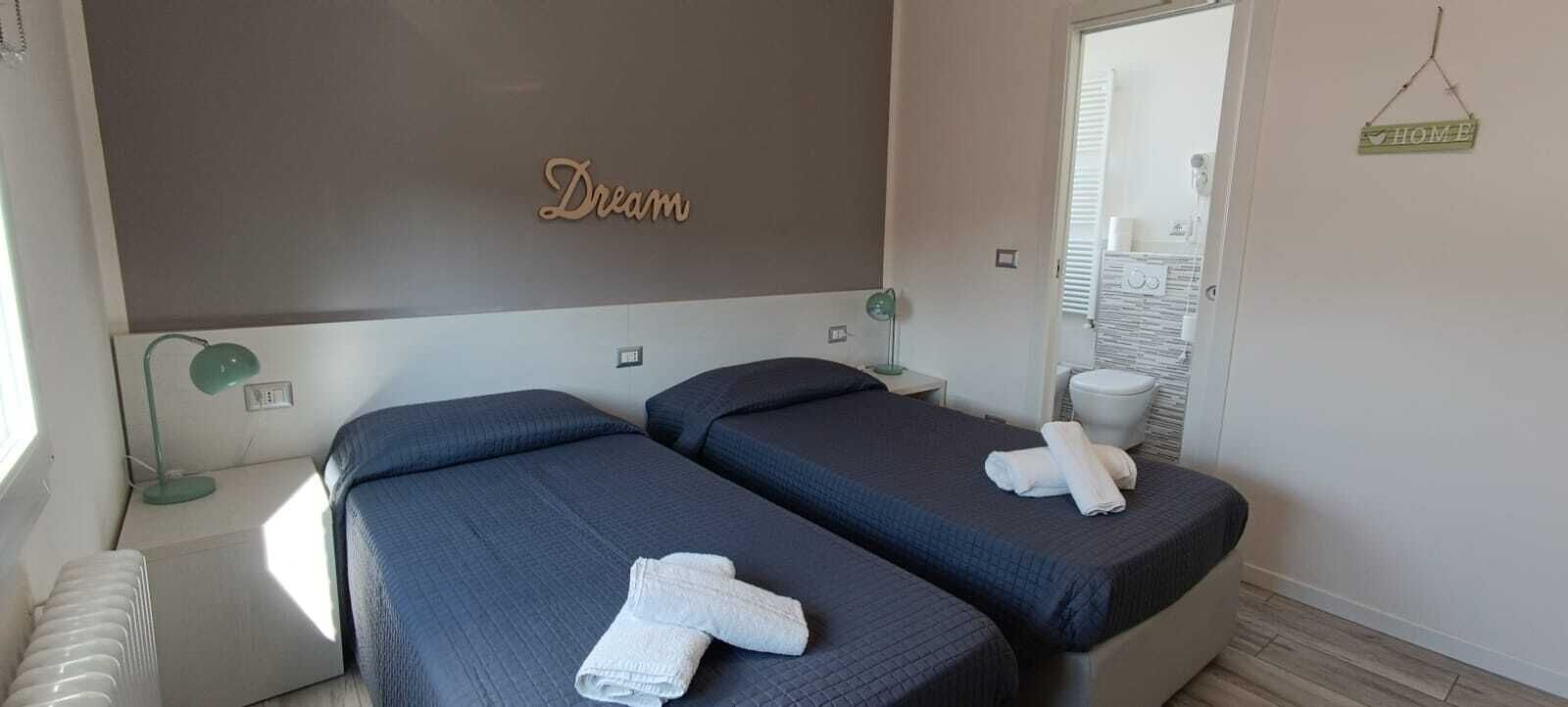A hotel room with two beds, a bedside table with a lamp, a mirror, and a 'Dream' sign on the wall.