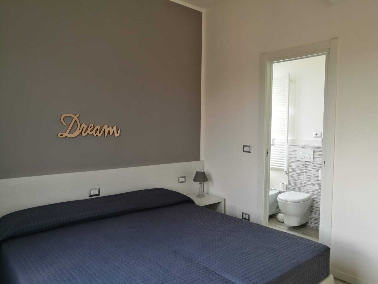Une chambre à coucher comprenant un lit, une table de chevet, une lampe, un panneau fixé au mur et donnant sur une salle de bain.