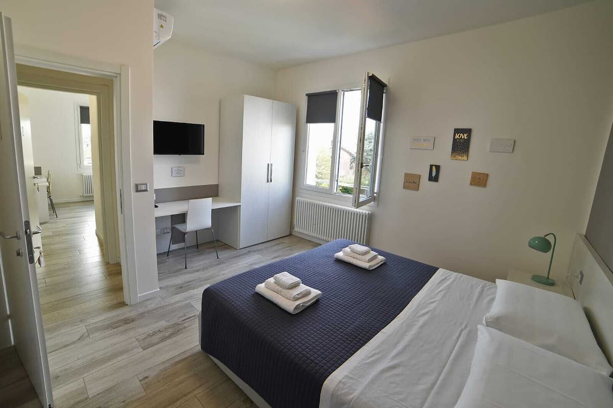 A bedroom with a bed, a desk, a wardrobe, a TV, and a window.