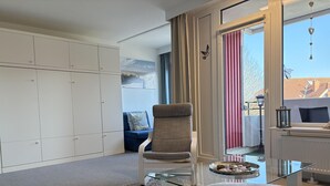 Una habitación moderna con una mesa de centro de vidrio, un sillón color beige, un sofá azul, un armario blanco y vistas al exterior a través de un ventanal grande.
