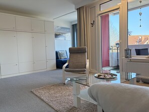 A modern room with a glass coffee table, a beige armchair, a blue sofa, a white wardrobe, and a view of the outdoors through a large window. - Berolina 112 - Lucy - 38 m² living space for max. 2 persons on the 1st floor (Dahme)