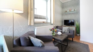 Un salon moderne avec un canapé, une table basse ornée d’un vase de fleurs, un téléviseur à écran plat et une fenêtre avec des stores.