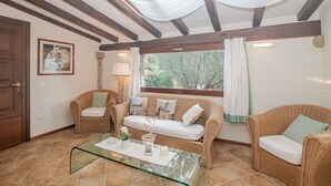 Una habitación con una mesa de centro de vidrio, sofás de mimbre, un cuadro enmarcado y una ventana con vistas a vegetación.