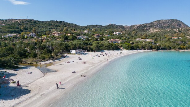 Una playa costera con agua turquesa cristalina, orilla arenosa y casas al fondo.