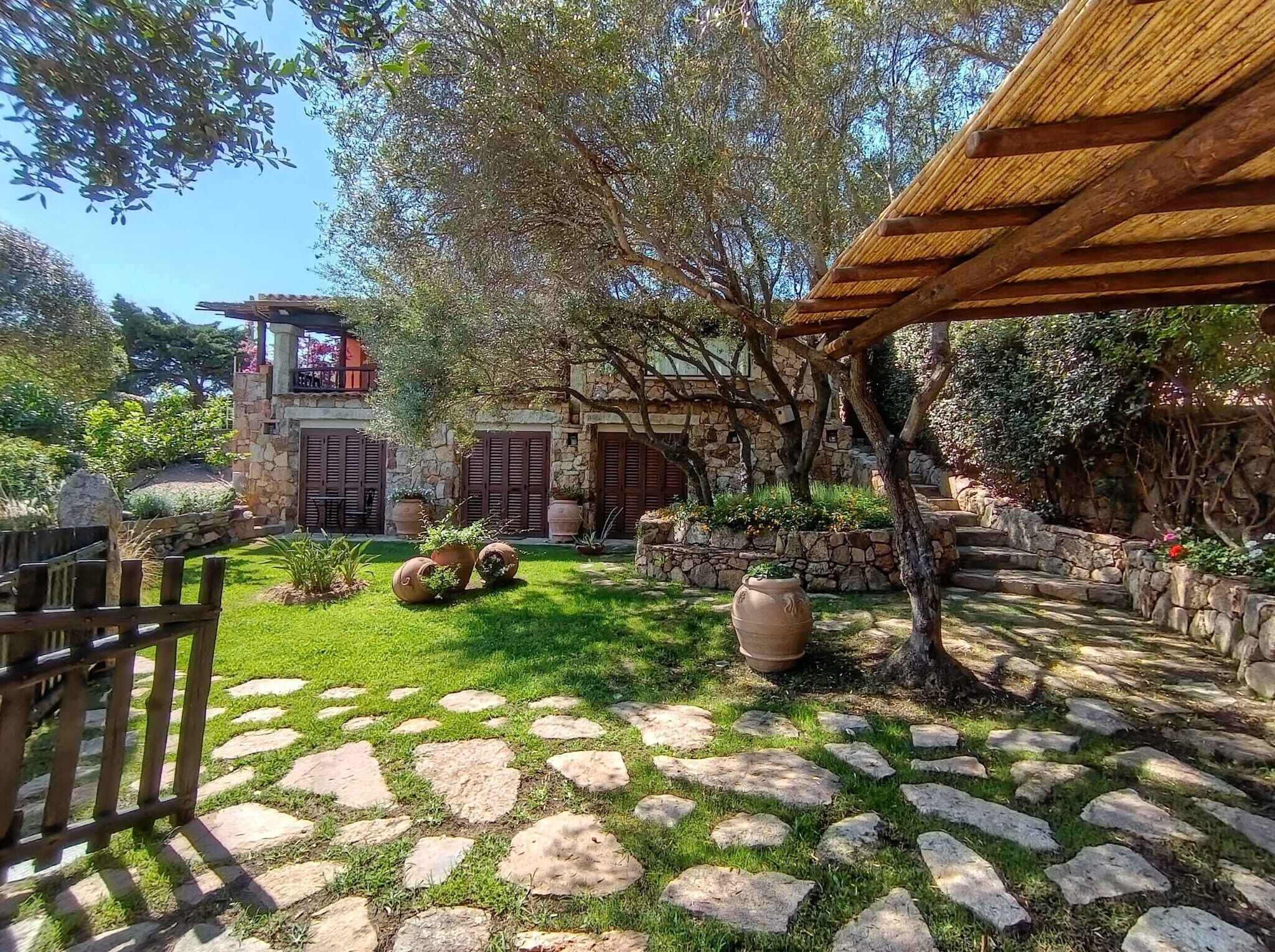 A stone building with a wooden pergola, a stone pathway, and a grassy area with potted plants.