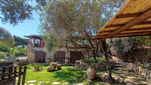 Un edificio de piedra con una pérgola de madera, un camino de piedra y una zona ajardinada con plantas en macetas.