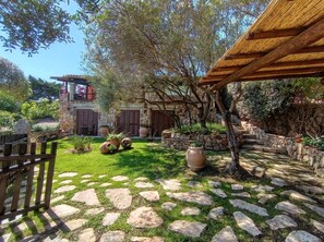 Un bâtiment en pierre agrémenté d’une pergola en bois, d’un chemin en pierre et d’une pelouse où sont disposées des plantes en pot.