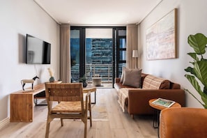 A modern living room with a sofa, a wooden desk, a TV, and a large window with a city view. - Eagle's Nest - Luxe 2BR with panoramic views, pool (Cape Town)