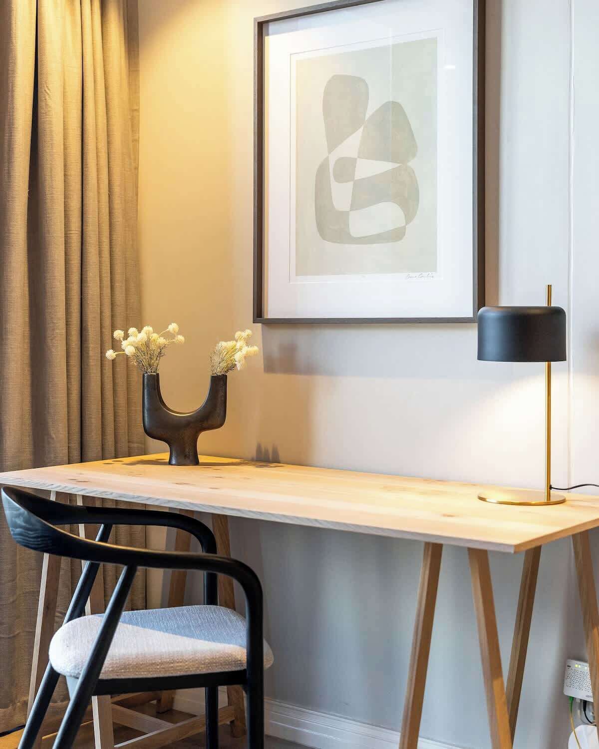 Un bureau moderne agrémenté d’un vase de fleurs séchées, d’une œuvre d’art abstraite encadrée, d’un lampadaire noir et d’une chaise.