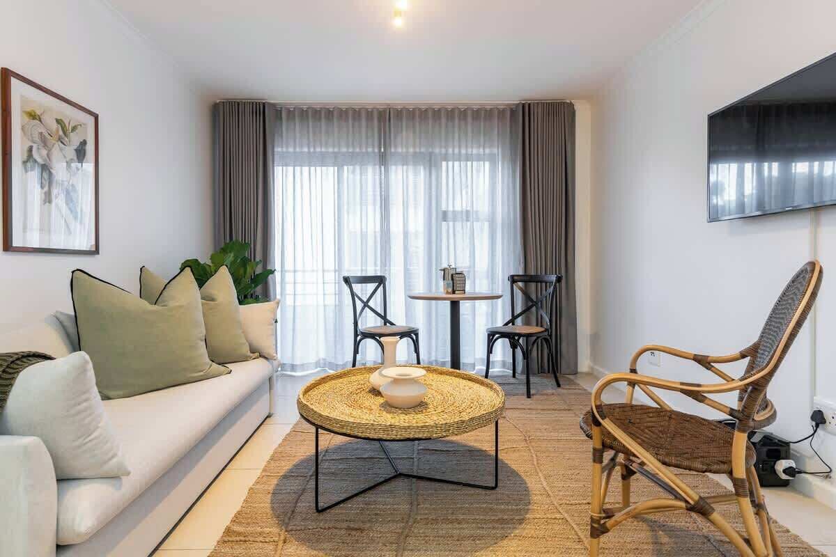 A modern living room with a sofa, a round coffee table, a wicker chair, and a TV mounted on the wall.