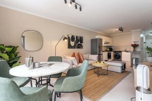 A modern living room with a round dining table, grey upholstered chairs, a sofa with cushions, a kitchen area with appliances, and a large mirror on the wall. - Mountain views - Designer 2BR, A/C, WiFi (Cape Town)