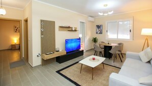 Una sala de estar moderna con televisor de pantalla plana, un comedor con sillas, un sofá blanco y una mesita con un objeto rojo.