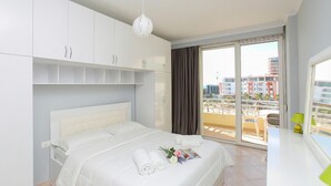 A modern hotel room with a bed, white cabinets, a balcony with a view of buildings, and a window with curtains. - Luna Sea View Apartment 5 Floor (Vlorë)