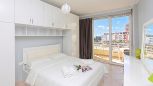 A modern hotel room with a bed, white cabinets, a large window with a view of buildings, and a balcony. - Luna Sea View Apartment 5 Floor (Vlorë)