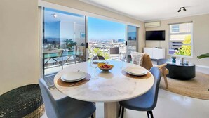 A modern living room with a dining area, large windows, and a flat-screen TV. - Casa del Mar: Sparkling secure 2BR, Wi-Fi, parking (Cape Town)