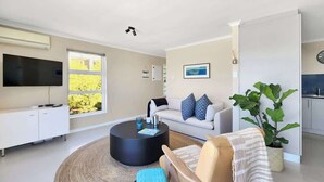 A modern living room with a flat-screen TV, a round coffee table, a sofa with patterned cushions, a chair with a striped throw, and a large plant. - Casa del Mar: Sparkling secure 2BR, Wi-Fi, parking (Cape Town)