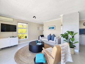 Ein modernes Wohnzimmer mit Flachbildfernseher, rundem Couchtisch, Sofa mit gemusterten Kissen, Sessel mit gestreifter Decke und einer großen Pflanze.