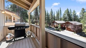 Um deck de madeira com churrasqueira e tanque de propano, com vista para uma cabana com garagem e rodeada por árvores.