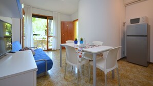 A compact room with a bed, a dining table with chairs, a TV, a refrigerator, and a window with curtains. - Perfect flat with garden and pool access (Bibione)