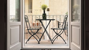 En åben balkon med et bord, stole, en potteplante og udsigt til en bygning med et vindue og en dør.